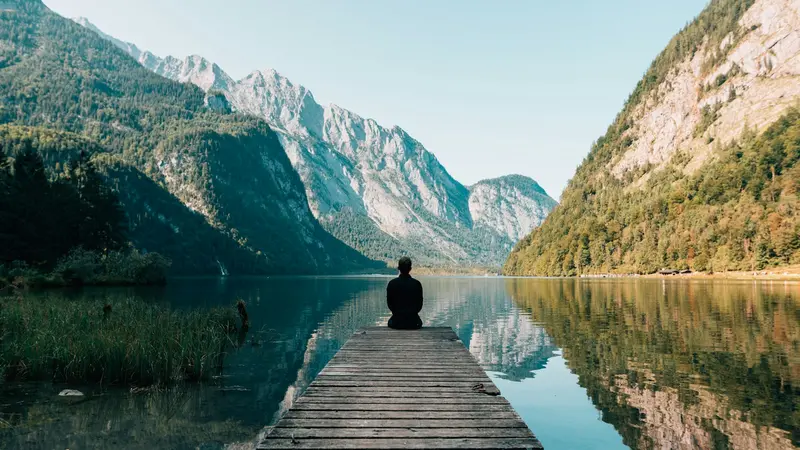 Person sitting at the end of a wooden dock on a calm mountain lake